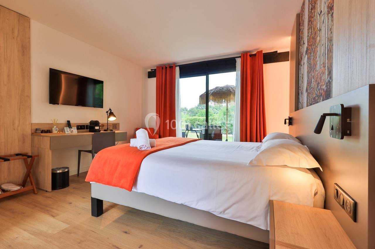 Chambre lumineuse avec lit double, draps blancs, plaid orange, bureau, télévision et vue sur un jardin par une baie vitrée.