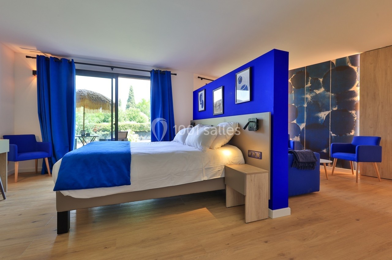 Chambre lumineuse avec lit double, décor bleu et blanc, grandes fenêtres donnant sur un jardin verdoyant.