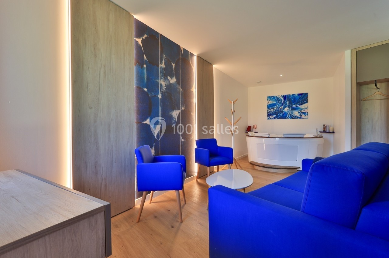 Salon moderne avec fauteuils bleus, table basse ronde, panneaux décoratifs et comptoir blanc dans un espace lumineux.