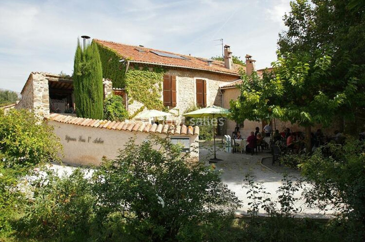 Façade d'une maison en pierre avec terrasse ombragée, parasols verts et végétation environnante.