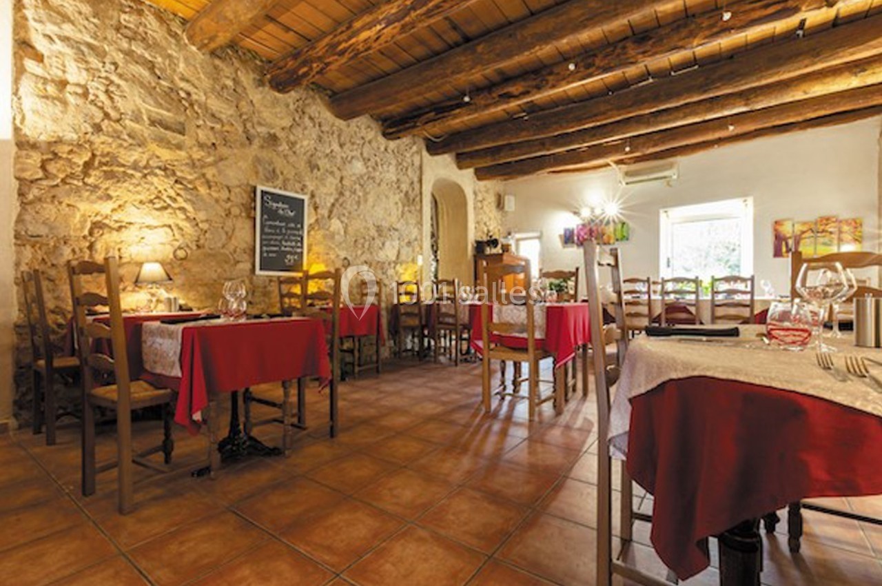 Salle de restaurant rustique avec murs en pierre, plafond en bois, tables dressées et nappes rouges.