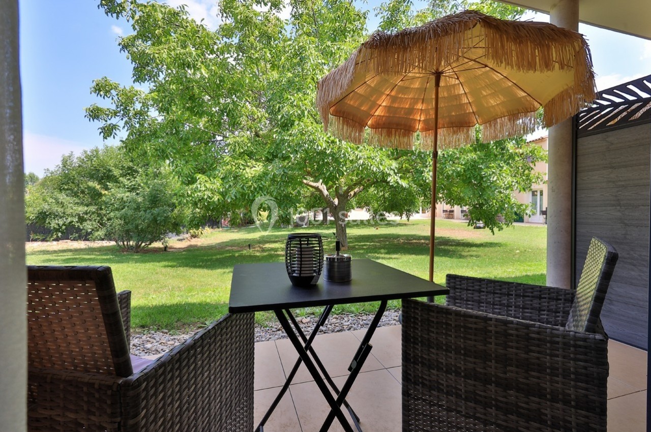 Table et chaises en rotin sous un parasol en paille, sur une terrasse avec vue sur un jardin verdoyant.