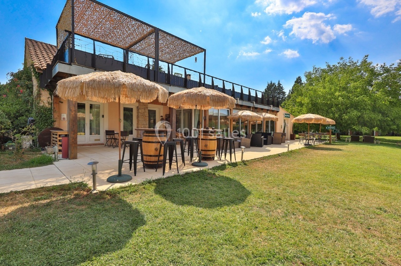 Terrasse d'un restaurant avec parasols en paille, tables et chaises, entourée de verdure sous un ciel ensoleillé.
