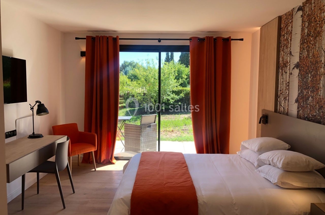 Chambre lumineuse avec lit double, bureau, chaise, rideaux orange et baie vitrée donnant sur un jardin verdoyant.