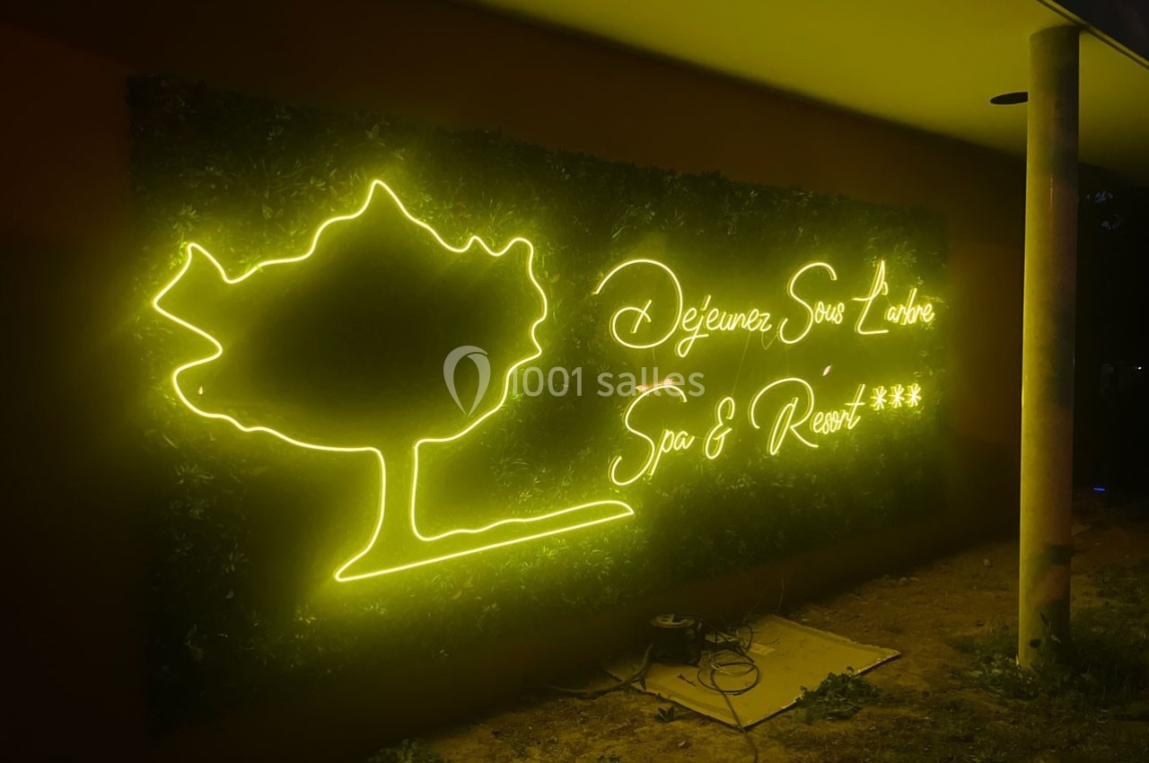 Enseigne lumineuse jaune représentant un arbre et le texte ’Déjeunez Sous l'Arbre Spa & Resort ***’ sur un mur sombre.