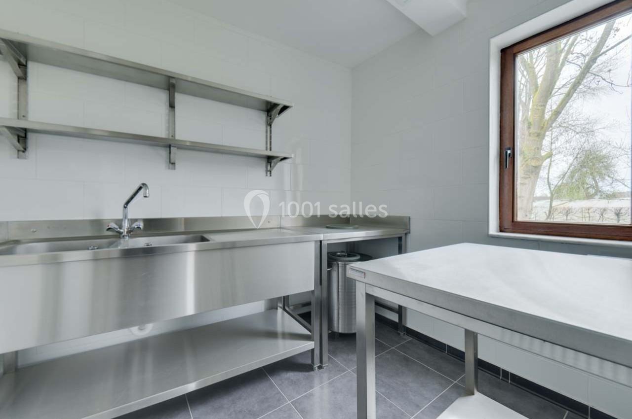 Cuisine professionnelle avec évier en inox, étagères murales, table de travail et fenêtre donnant sur l'extérieur.