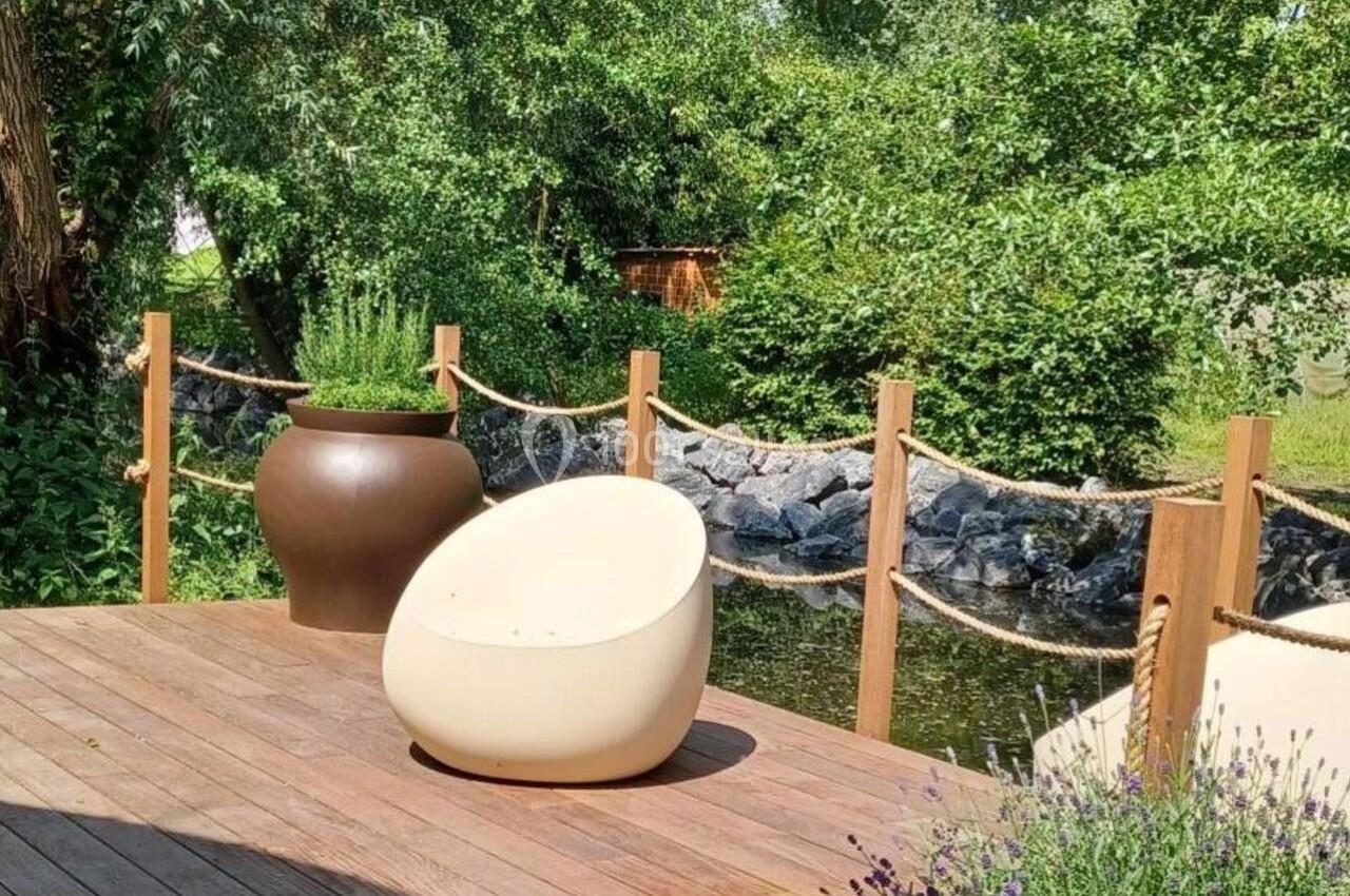 Fauteuil beige sur une terrasse en bois près d'un étang entouré de verdure et de plantes.