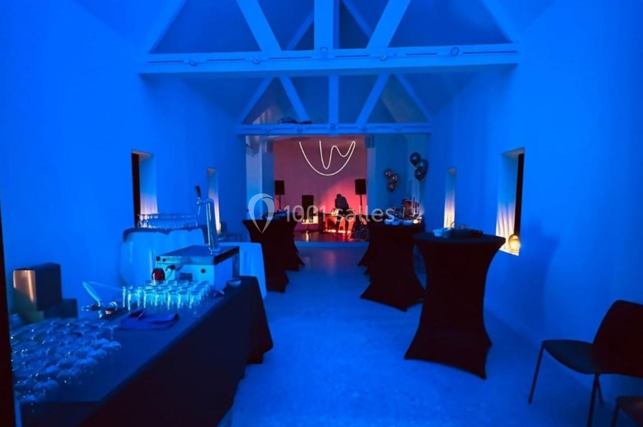 Salle éclairée en bleu avec tables hautes noires, buffet à gauche et ambiance lumineuse rouge au fond.