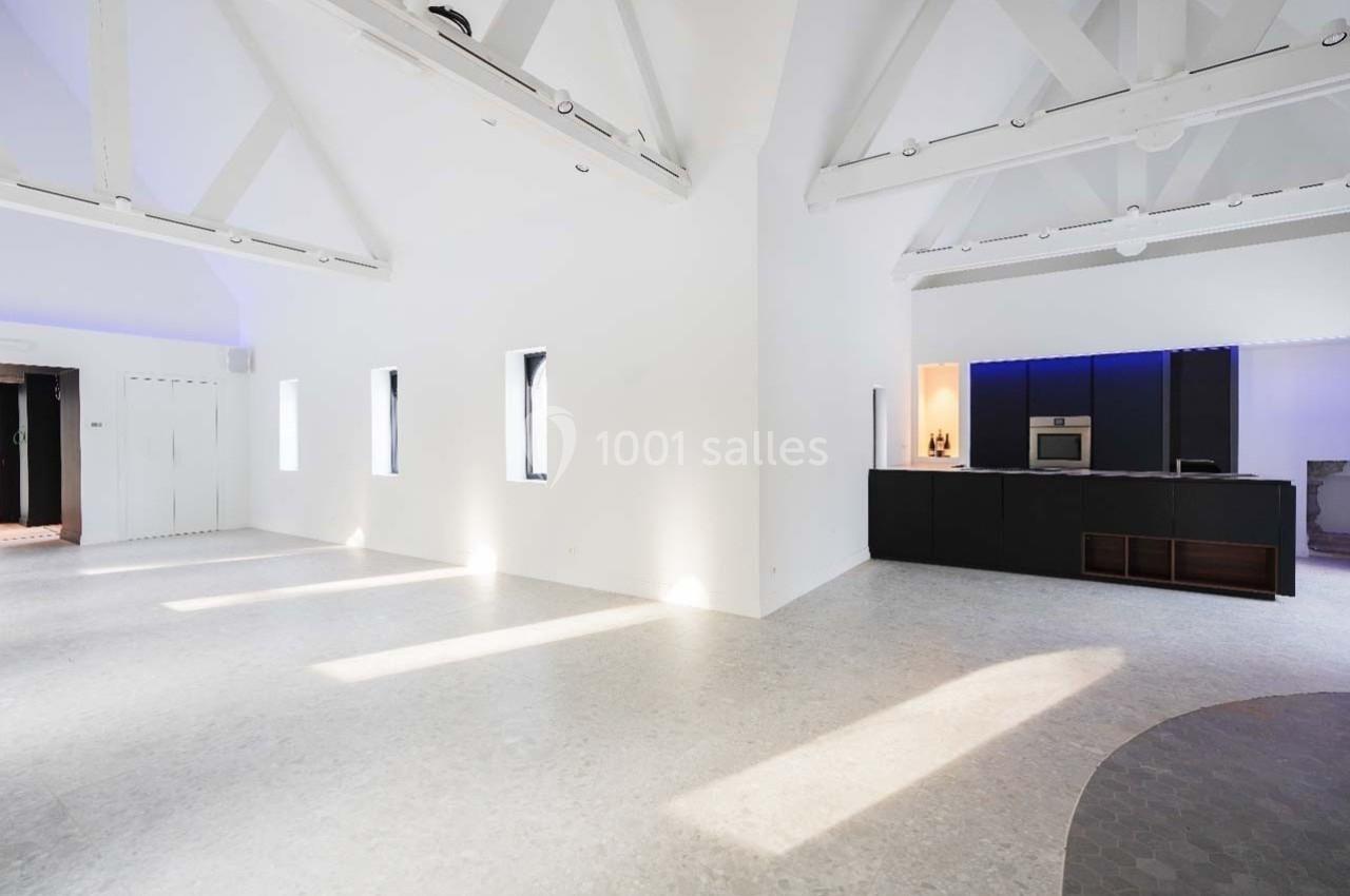 Intérieur lumineux avec plafond haut, poutres apparentes, sol clair et cuisine moderne avec éclairage intégré.