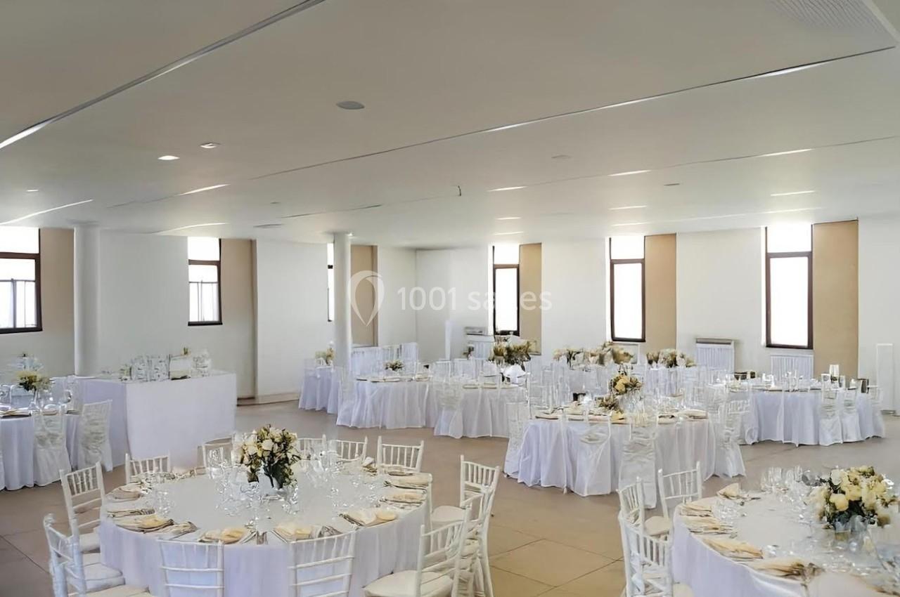 Salle de réception lumineuse avec tables rondes dressées, nappes blanches et décorations florales élégantes.