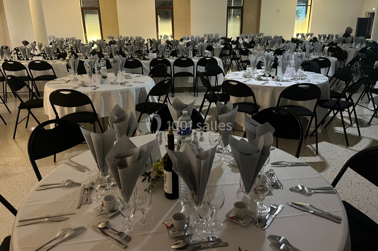 Salle de réception avec des tables rondes dressées, nappes blanches, serviettes pliées et couverts disposés.