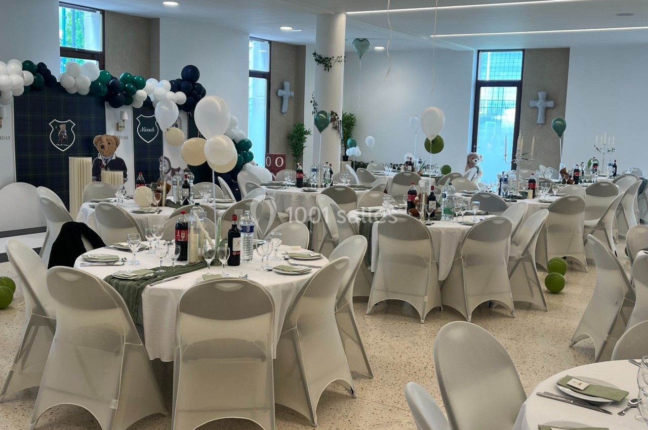 Salle décorée pour un événement avec des tables rondes dressées, des ballons et des éléments festifs.