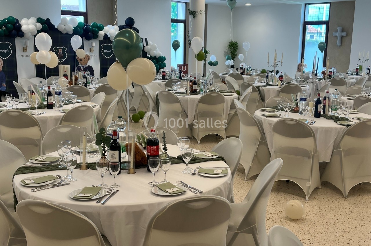 Salle décorée pour une réception avec des tables rondes dressées, ballons et décorations en arrière-plan.