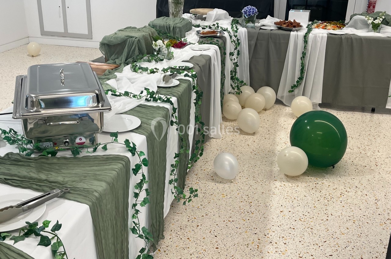 Buffet décoré de nappes blanches et vertes avec des guirlandes végétales, entouré de ballons blancs et verts.