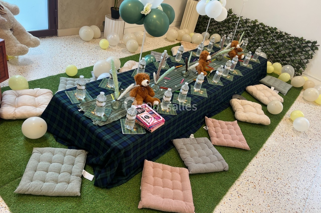 Table basse décorée pour une fête d'enfants, avec peluches, ballons, coussins et bouteilles d'eau sur une nappe à carreaux.