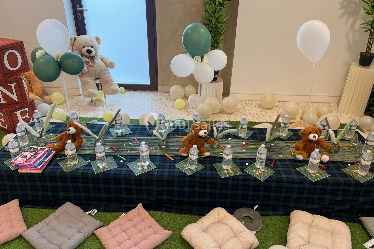 Table basse décorée pour une fête d'enfants avec peluches, ballons, coussins et bouteilles d'eau alignées.