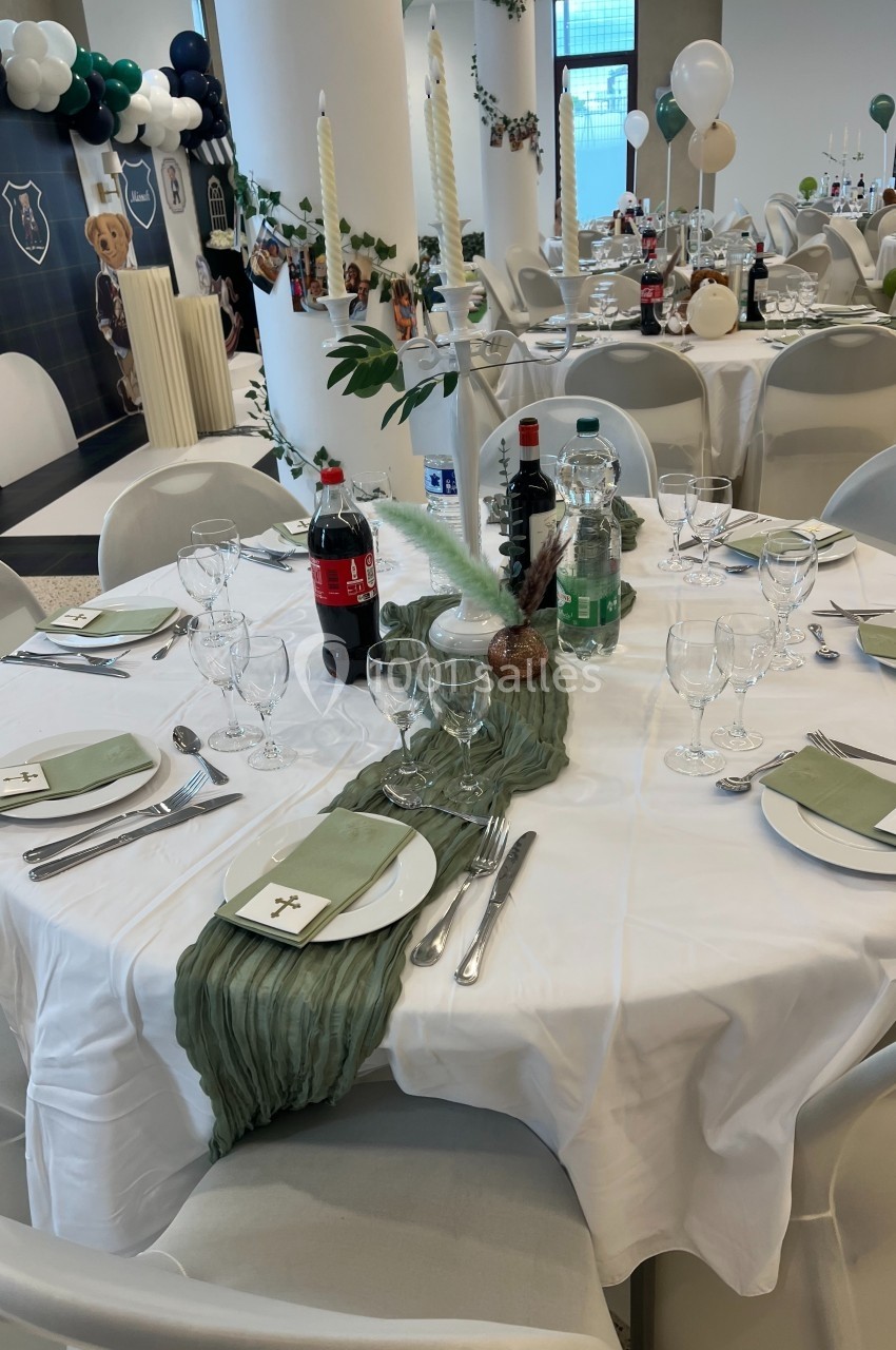 Table ronde dressée pour un événement, avec nappes blanches, serviettes vertes, bougies et boissons disposées au centre.
