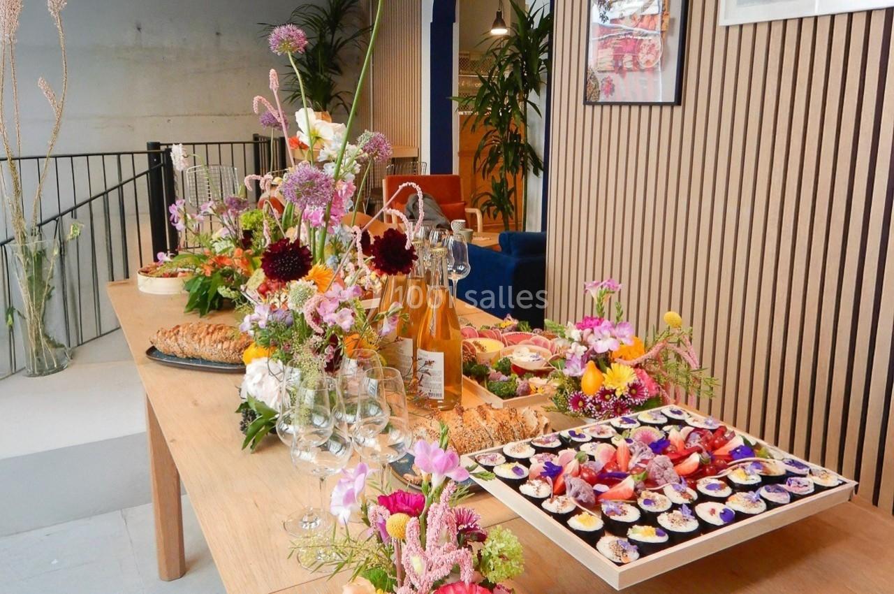 Table en bois garnie de fleurs colorées, plats variés, bouteilles de vin et verres dans un espace moderne et lumineux.