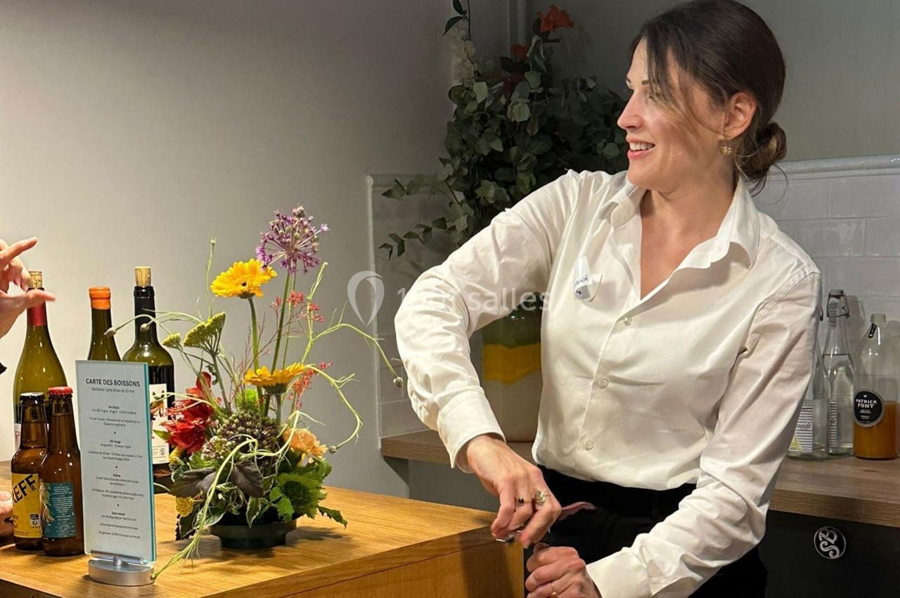 Une femme en chemise blanche ouvre une bouteille derrière un comptoir décoré de fleurs et de boissons.