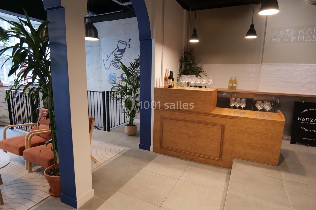 Espace intérieur moderne avec comptoir en bois, étagères à verres, plantes et coin salon lumineux.