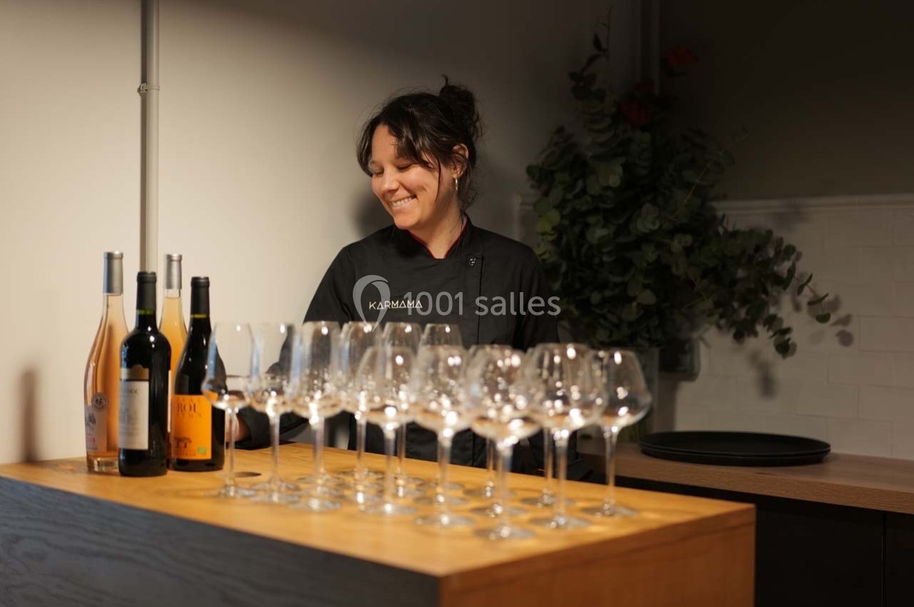 Une femme souriante derrière un comptoir avec des verres à vin alignés et des bouteilles de vin.