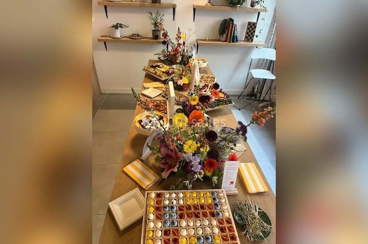 Table garnie de fleurs colorées, pâtisseries variées et chocolats, dans un espace lumineux avec étagères en arrière-plan.
