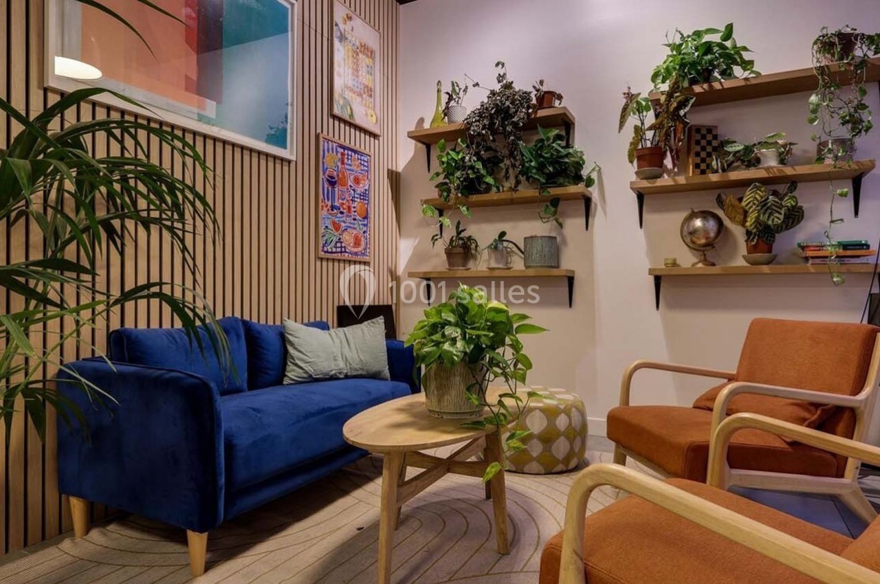 Salon moderne avec canapé bleu, fauteuils orange, plantes vertes sur étagères et décoration murale colorée.