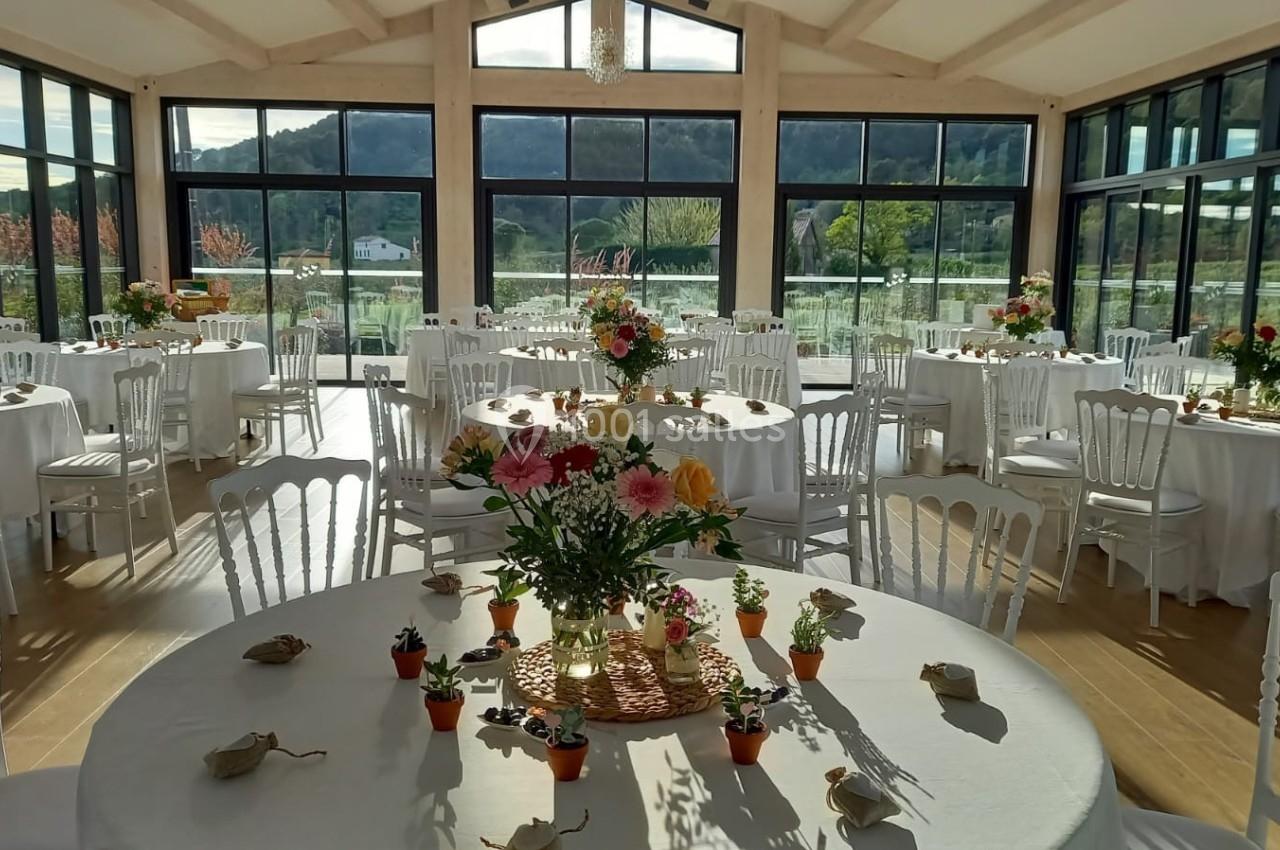Salle lumineuse avec grandes baies vitrées, tables rondes décorées de fleurs et petites plantes, vue sur un paysage…