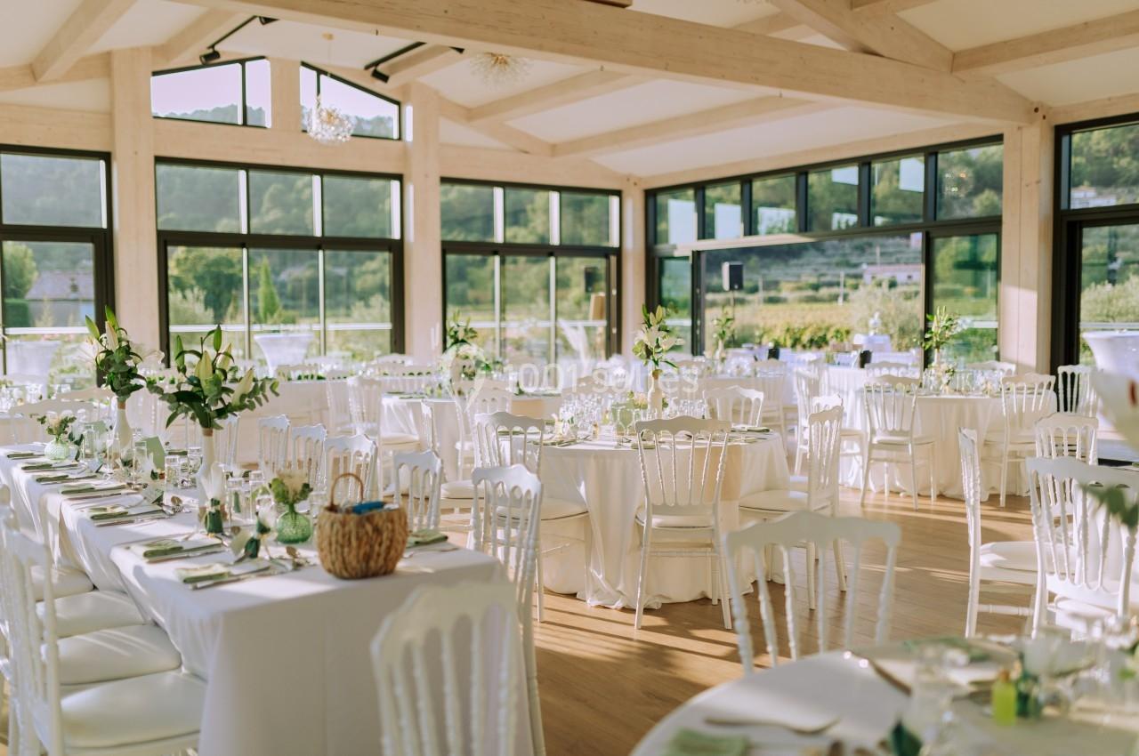 Salle lumineuse décorée pour un événement, avec tables dressées, chaises blanches et grandes baies vitrées donnant sur un…