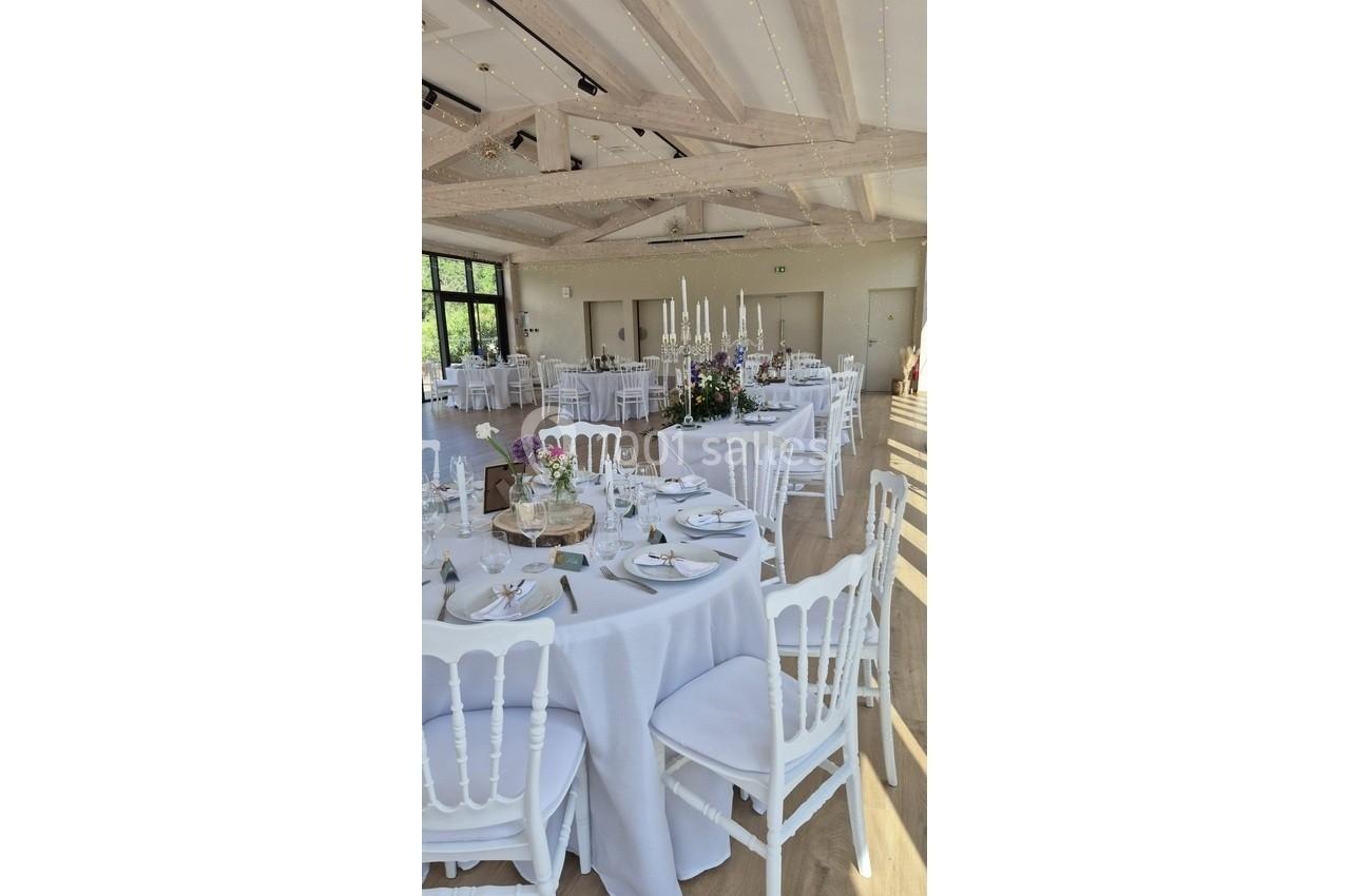 Salle lumineuse décorée pour un événement, tables rondes dressées avec nappes blanches, chaises et centre de table floral.