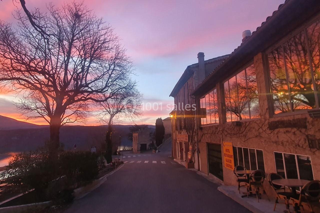 Rue bordée de bâtiments en pierre et d'arbres au coucher du soleil, avec des reflets roses dans le ciel.