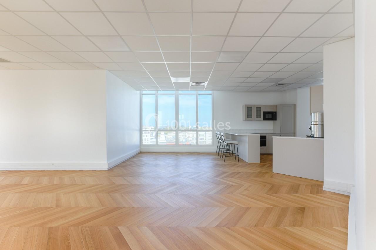 Salle lumineuse avec parquet en chevrons, grande fenêtre, cuisine ouverte et plafond à dalles blanches.