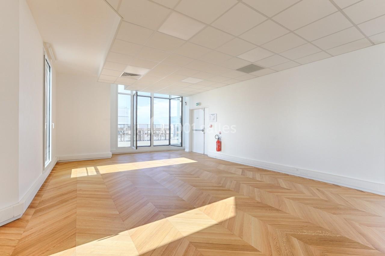 Salle lumineuse avec parquet en chevrons, grandes baies vitrées donnant sur une terrasse et plafond à dalles blanches.