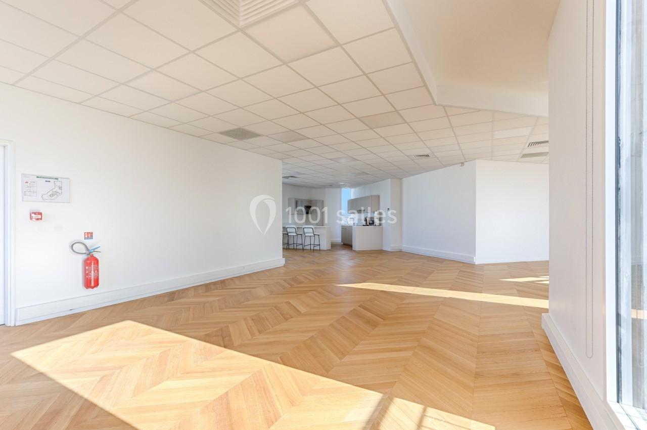 Salle lumineuse avec parquet en chevrons, murs blancs, coin cuisine au fond et extincteur fixé au mur.