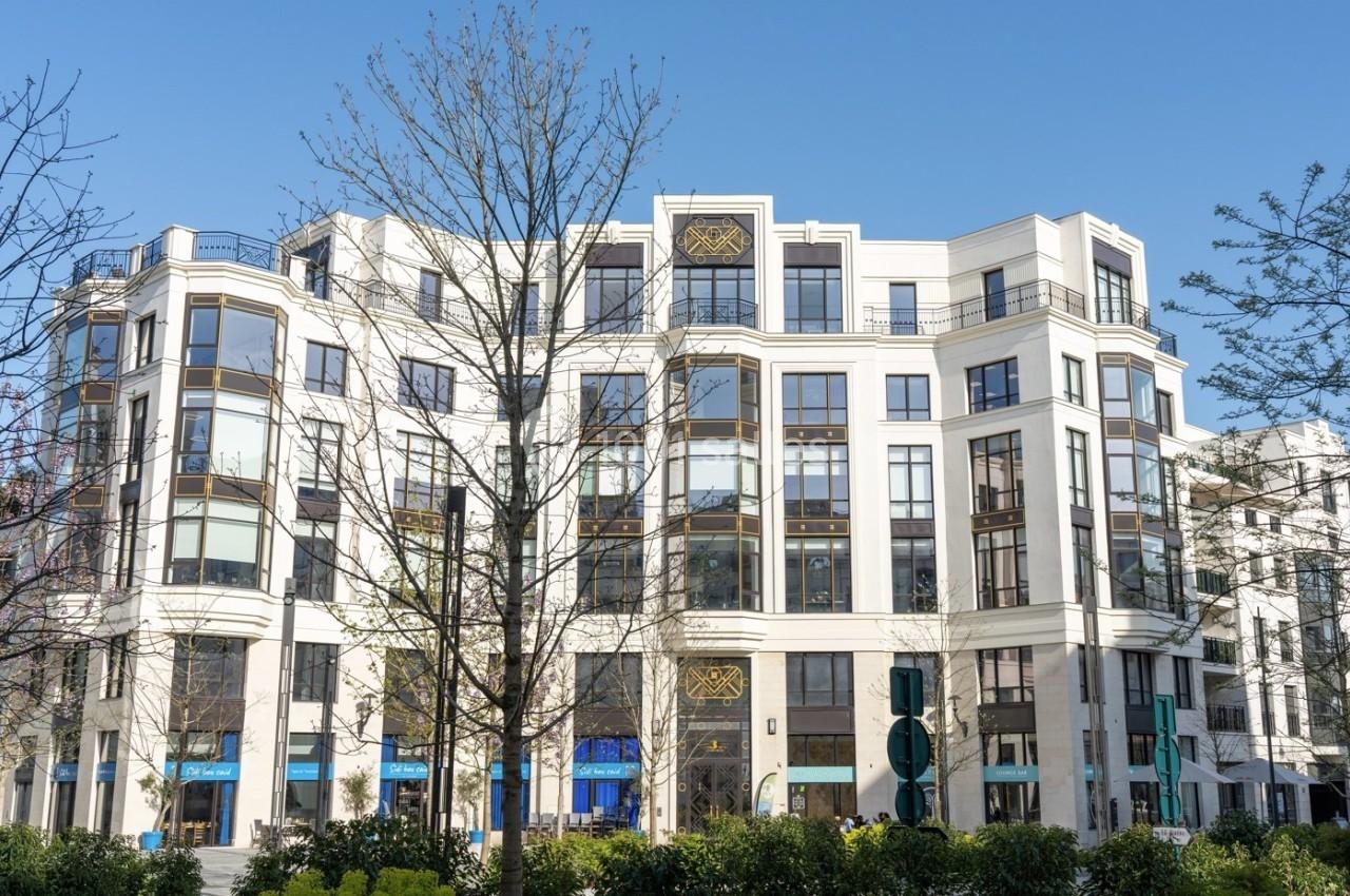 Bâtiment moderne à plusieurs étages avec des façades blanches, de grandes fenêtres et des arbres au premier plan.
