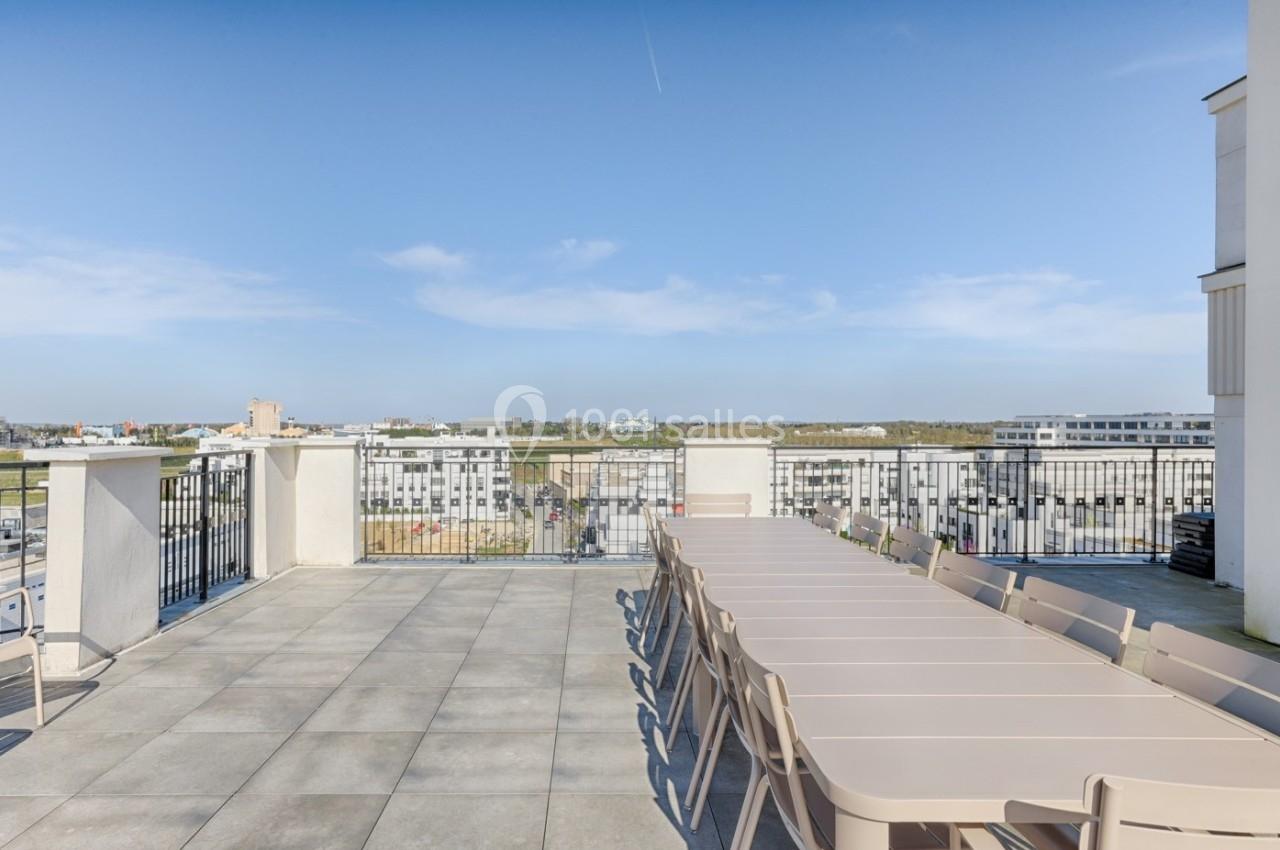 Terrasse spacieuse avec grande table et chaises, offrant une vue dégagée sur des bâtiments et un horizon lointain.