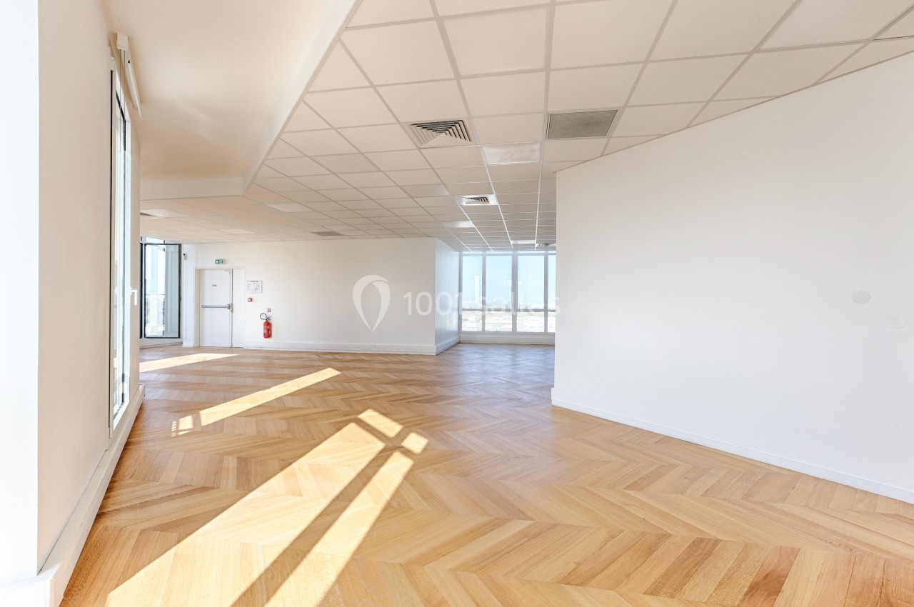 Bureau vide avec parquet en chevrons, murs blancs, plafond à dalles et grandes fenêtres laissant entrer la lumière naturelle.