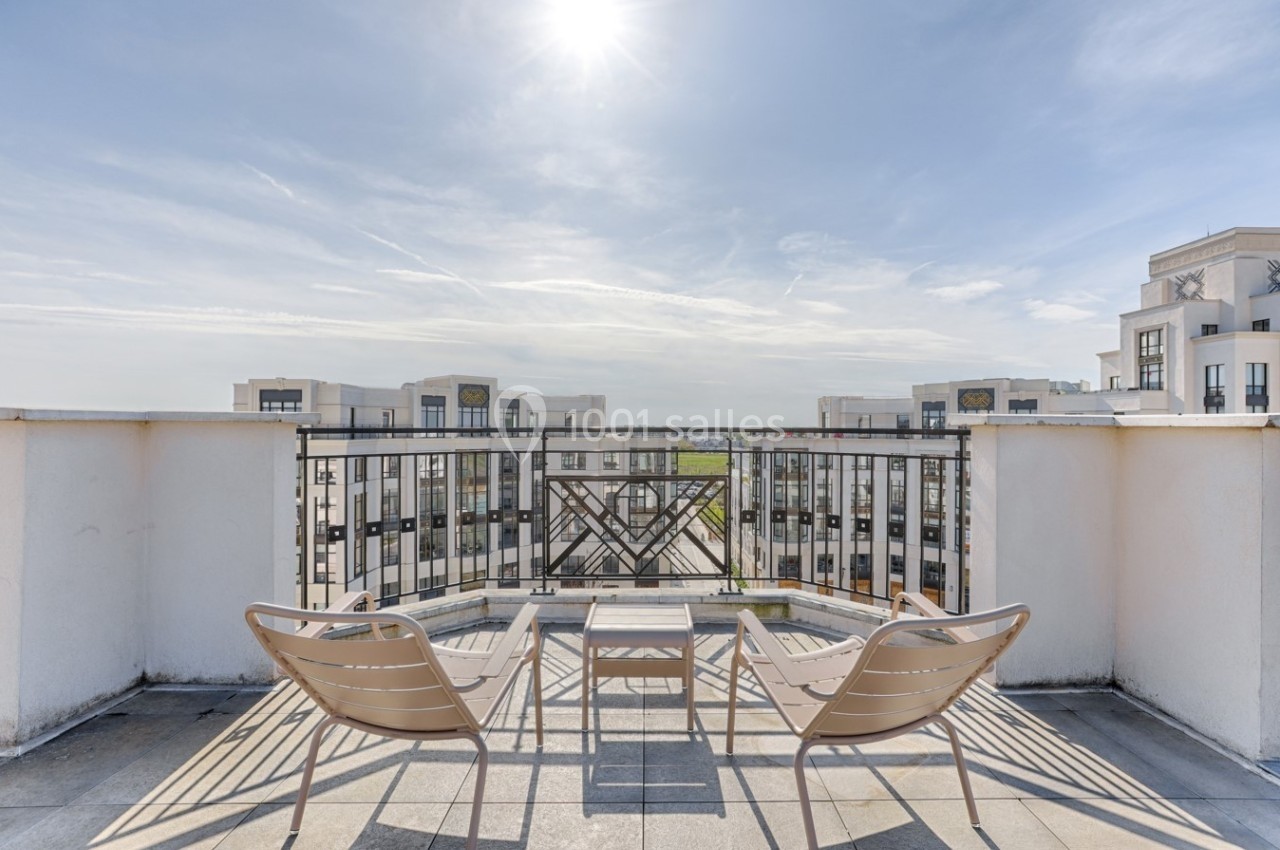 Deux chaises et une table sur une terrasse ensoleillée avec vue sur des immeubles modernes et un ciel dégagé.