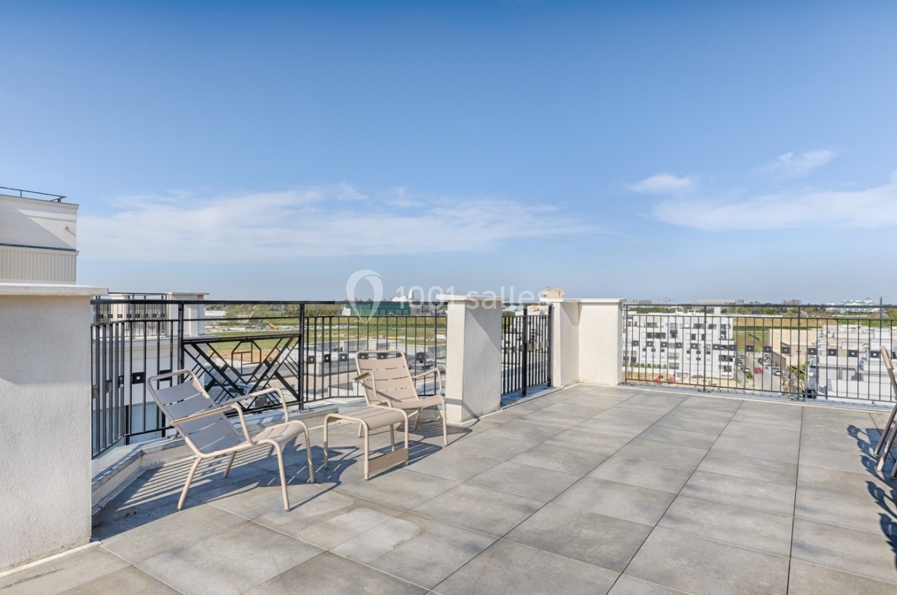 Terrasse spacieuse avec sol en carrelage, chaises longues et vue dégagée sur des bâtiments et un ciel bleu.