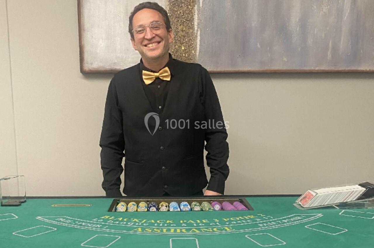 Un croupier souriant en tenue formelle se tient derrière une table de blackjack avec des jetons et des cartes.