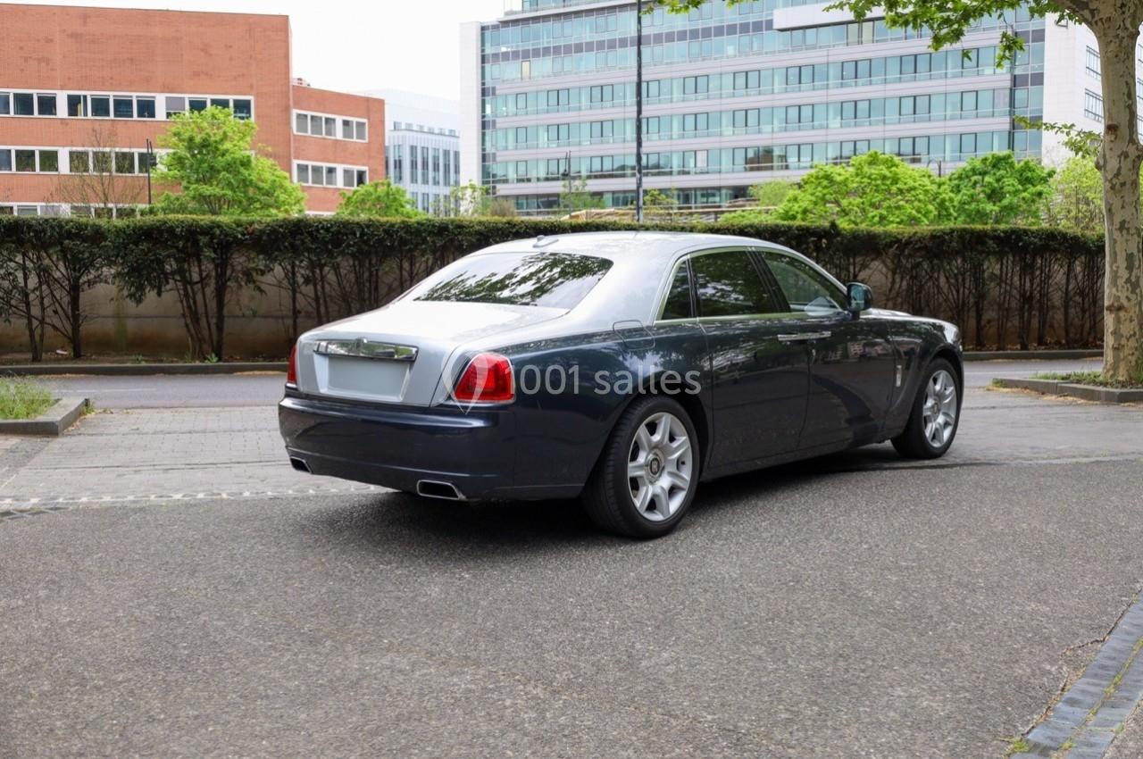 Voiture de luxe sombre stationnée sur une route bordée d'arbres, avec des bâtiments modernes en arrière-plan.