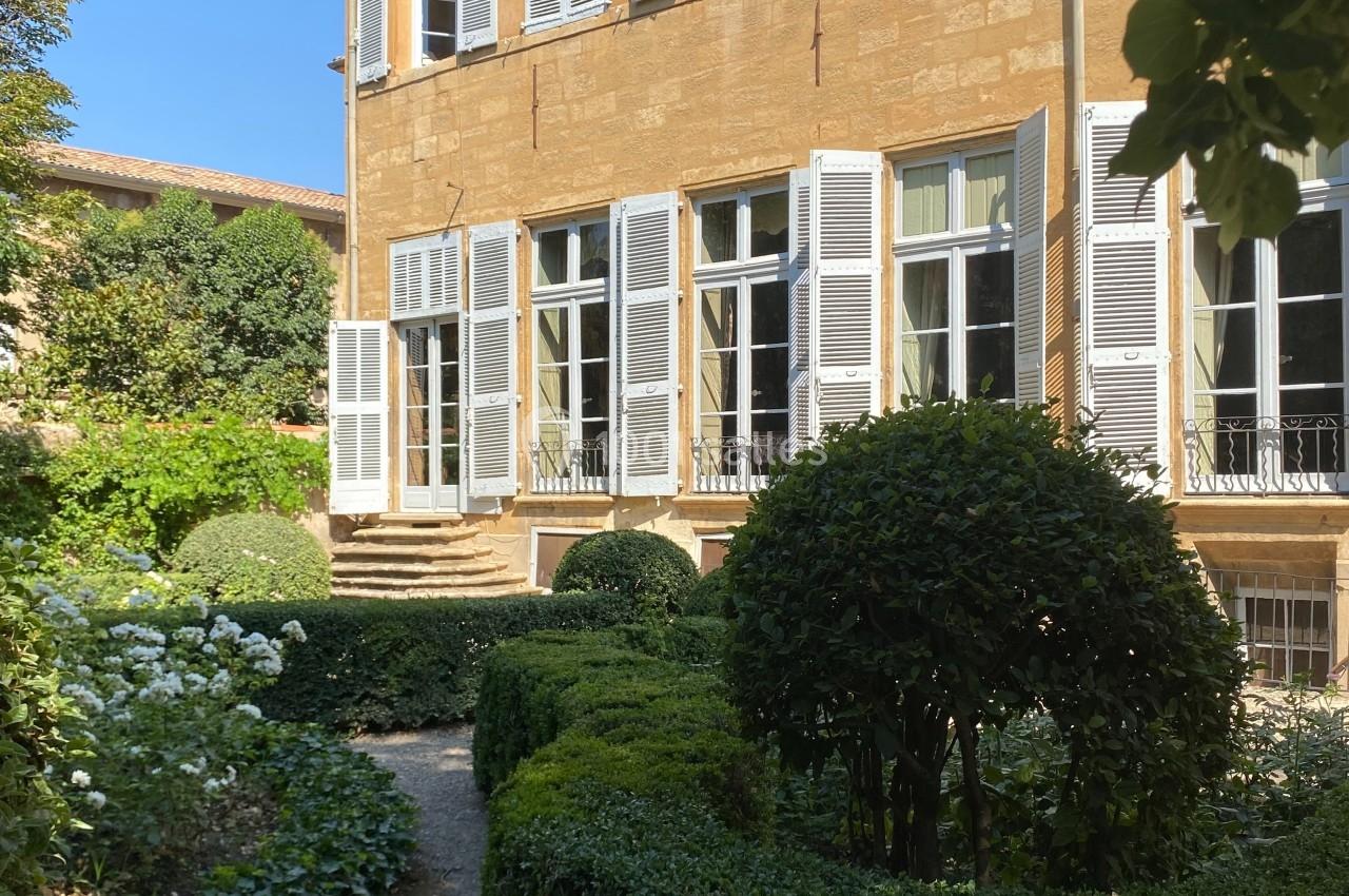 Façade d'une maison en pierre ocre avec volets blancs, entourée d'un jardin verdoyant et bien entretenu.