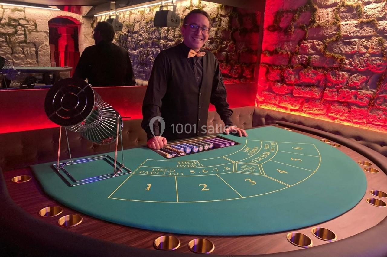 Homme souriant debout derrière une table de jeu de casino avec un cylindre de tirage et des jetons alignés.