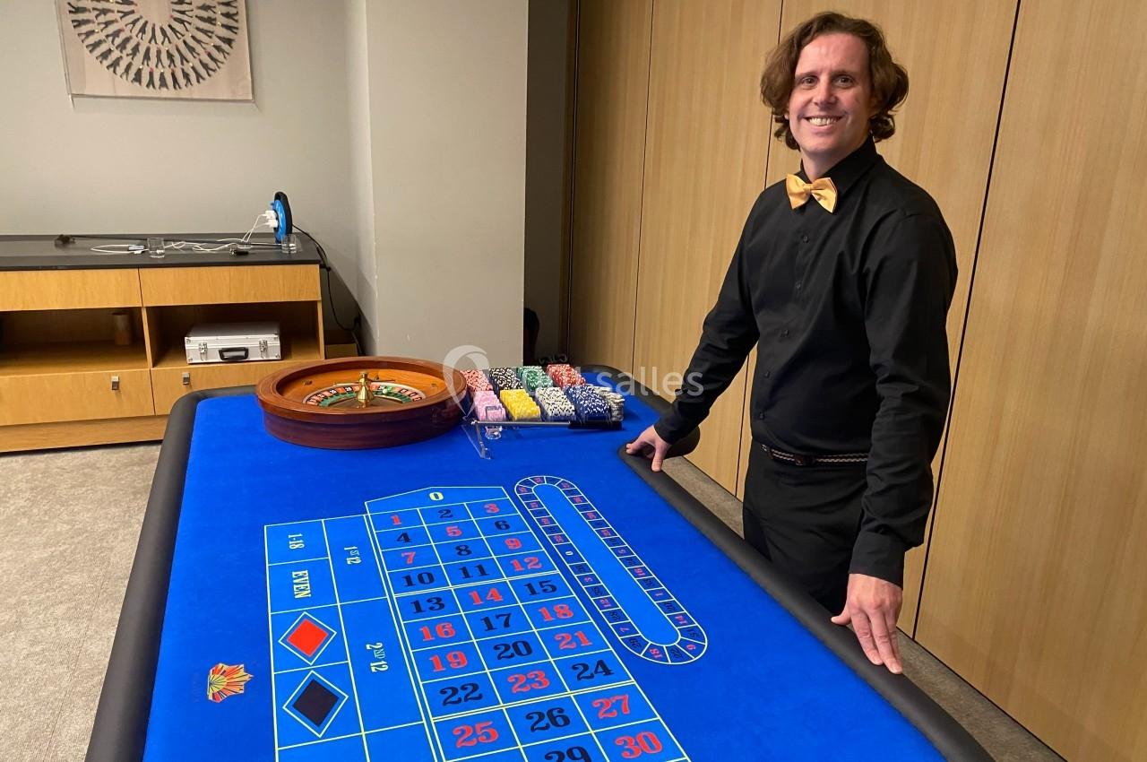 Homme souriant debout près d'une table de roulette avec jetons colorés et roue de jeu dans une pièce sobre.