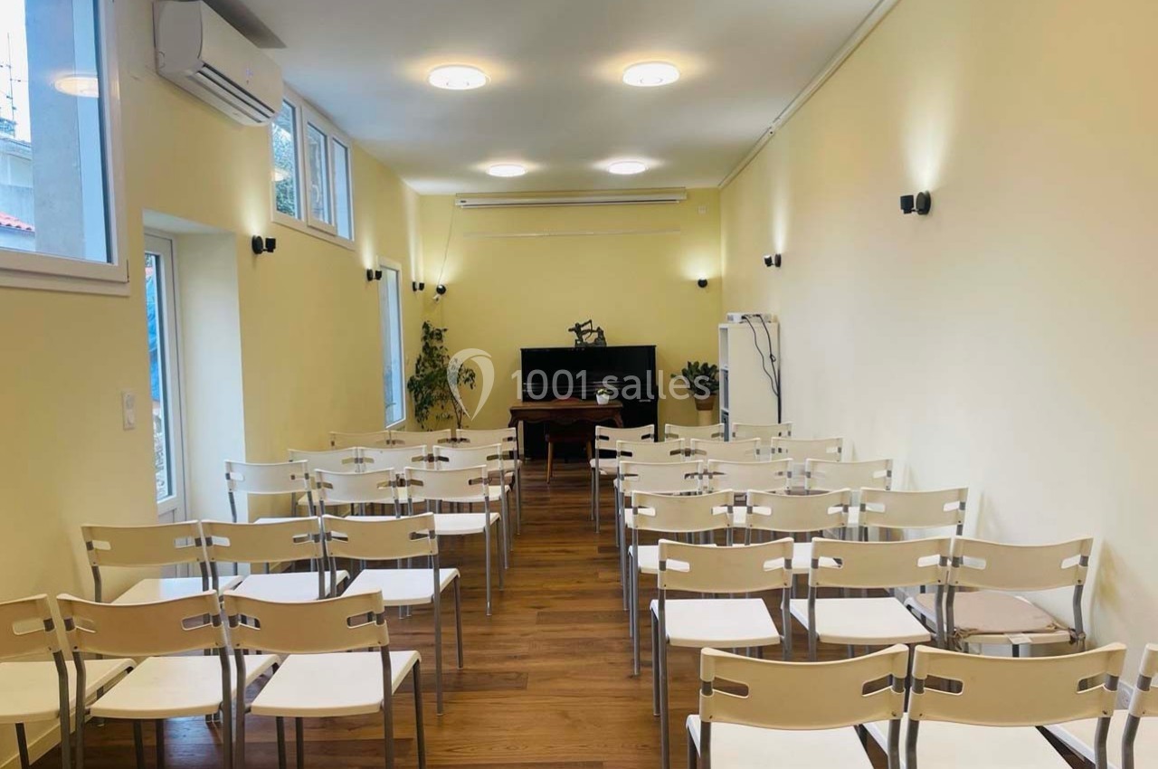 Salle lumineuse avec des rangées de chaises blanches, un bureau et un écran au fond, parquet en bois au sol.
