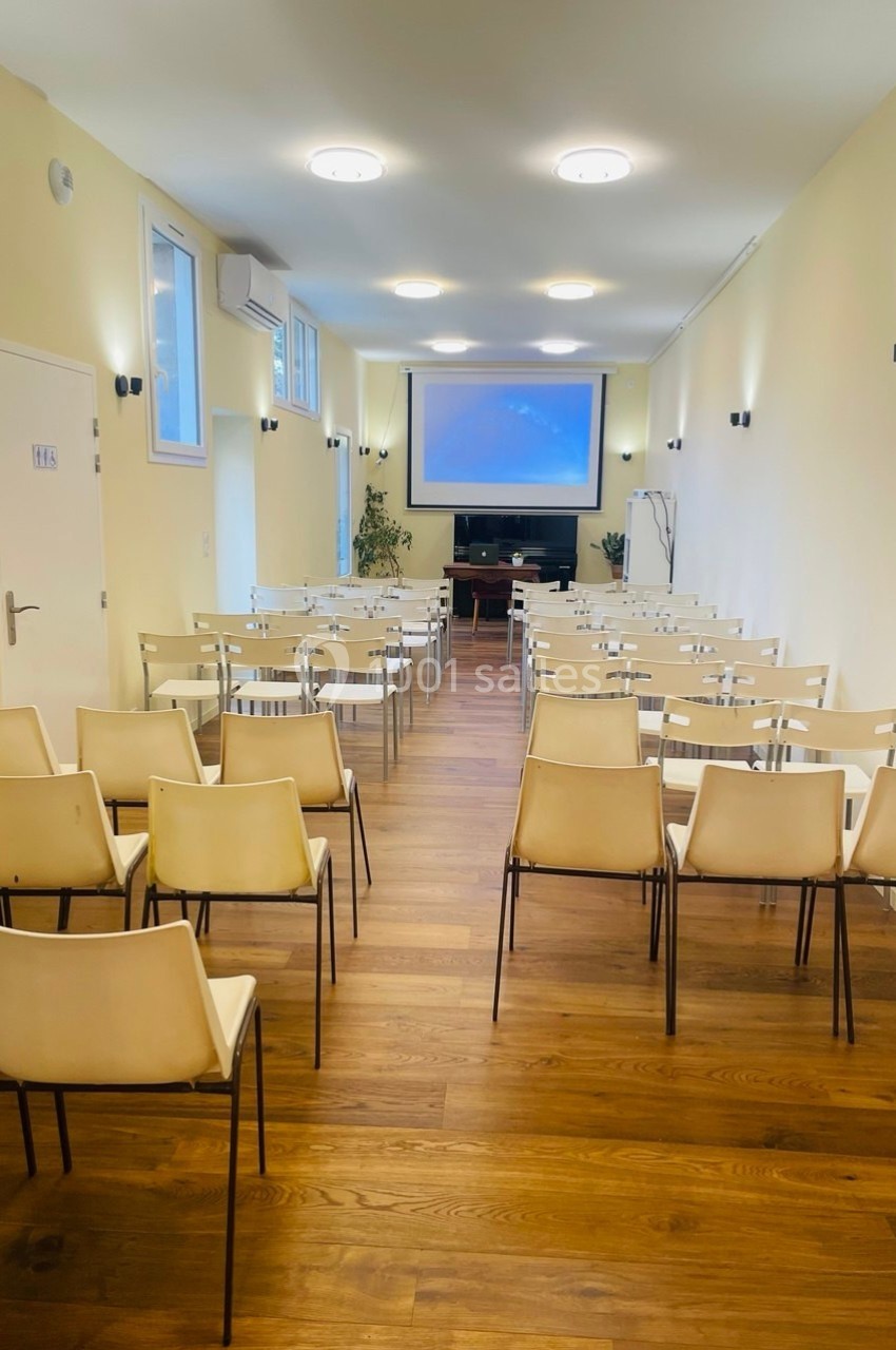 Salle lumineuse avec rangées de chaises alignées face à un écran de projection, parquet en bois au sol.