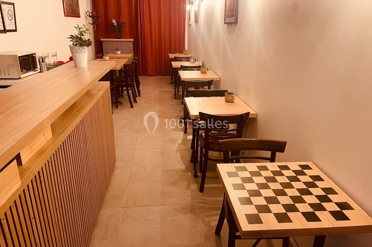 Salle de café avec tables en bois, tabourets, échiquier au premier plan et rideaux rouges en arrière-plan.