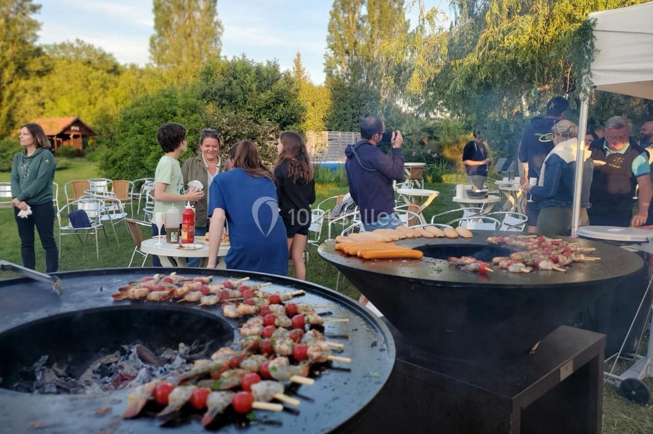 Personnes discutant près de barbecues en plein air, avec brochettes et saucisses en cours de cuisson.