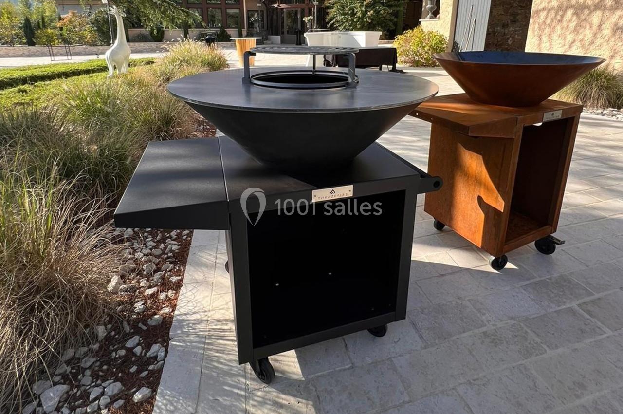 Plancha ronde sur chariot noir avec tablette latérale, exposée en extérieur sur une terrasse pavée.