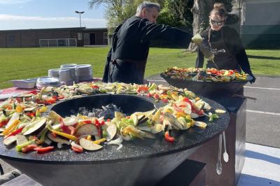 Table de pique-nique avec nappe à carreaux, paniers, pain, huîtres et vin près d'un brasero allumé dans un jardin.